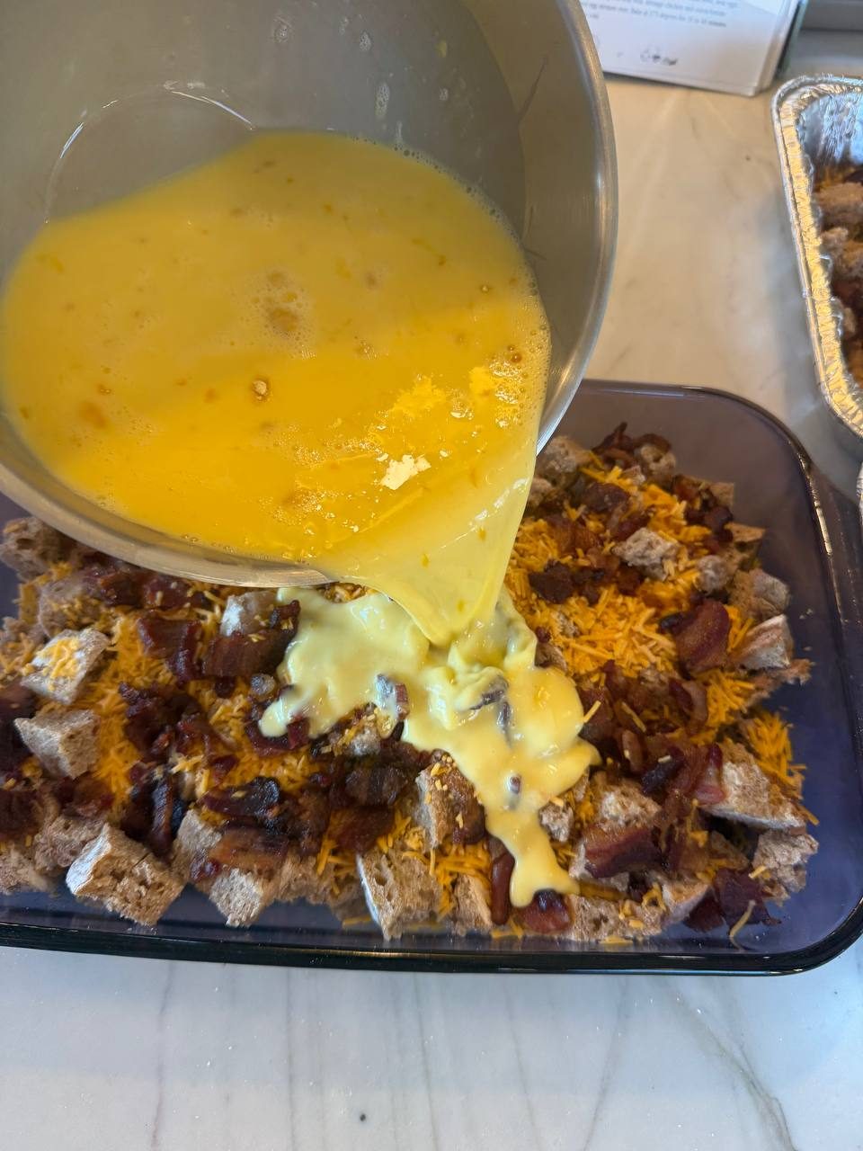 Beaten eggs pouring over bread, cheddar, and bacon in a 9x13 dish